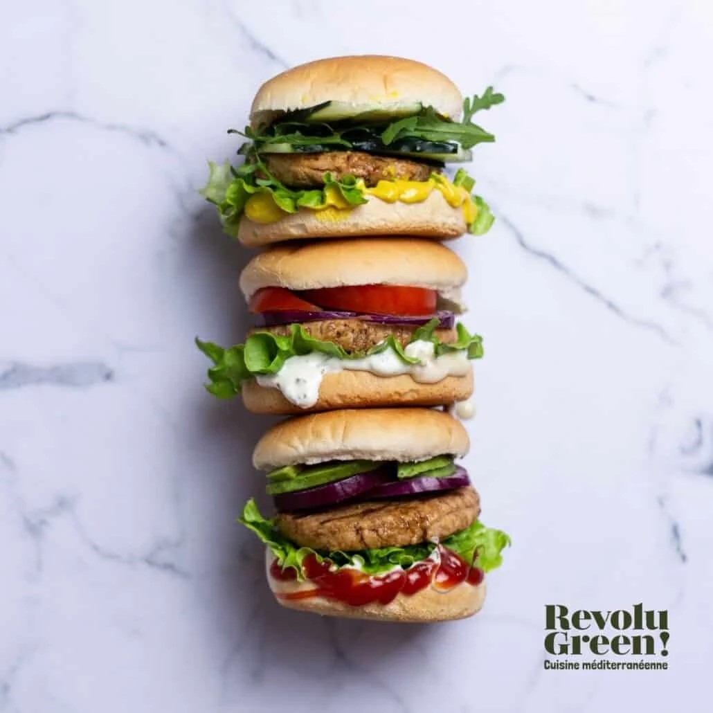A tall, vertical stack of three plant-based sliders arranged on a white marble surface. Each burger features unique toppings including leafy greens, sliced red onions, cucumbers, tomatoes, avocado slices, and various condiments. The bottom right corner displays a logo that reads "Revolu Green! Cuisine méditerranéenne".