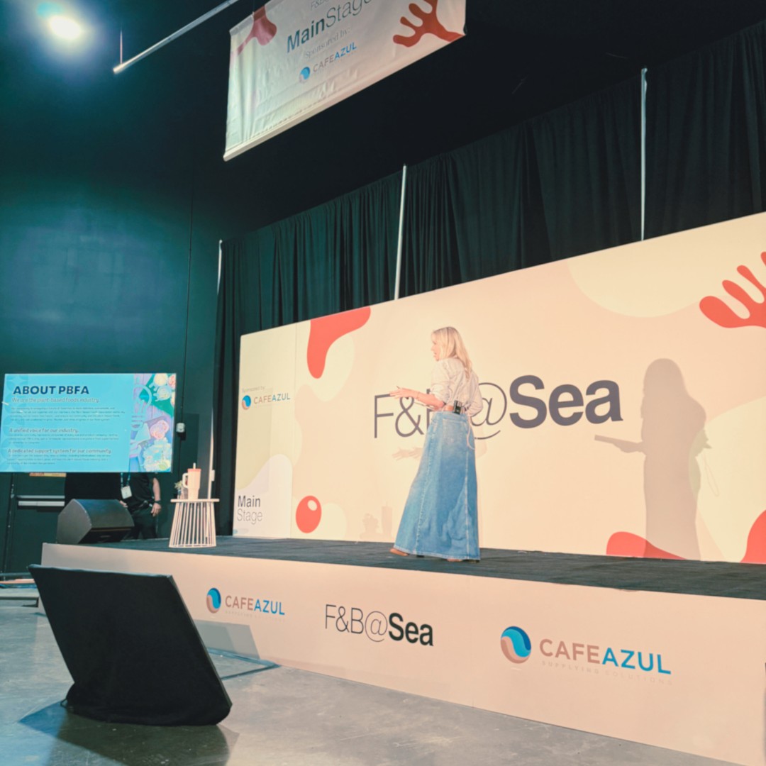 A speaker with long blonde hair, wearing a striped shirt and a long denim maxi skirt, stands on a convention stage presenting at the "F&B@Sea" event. The stage features banners and signage for "CAFEAZUL" and a side monitor displaying information about PBFA.
