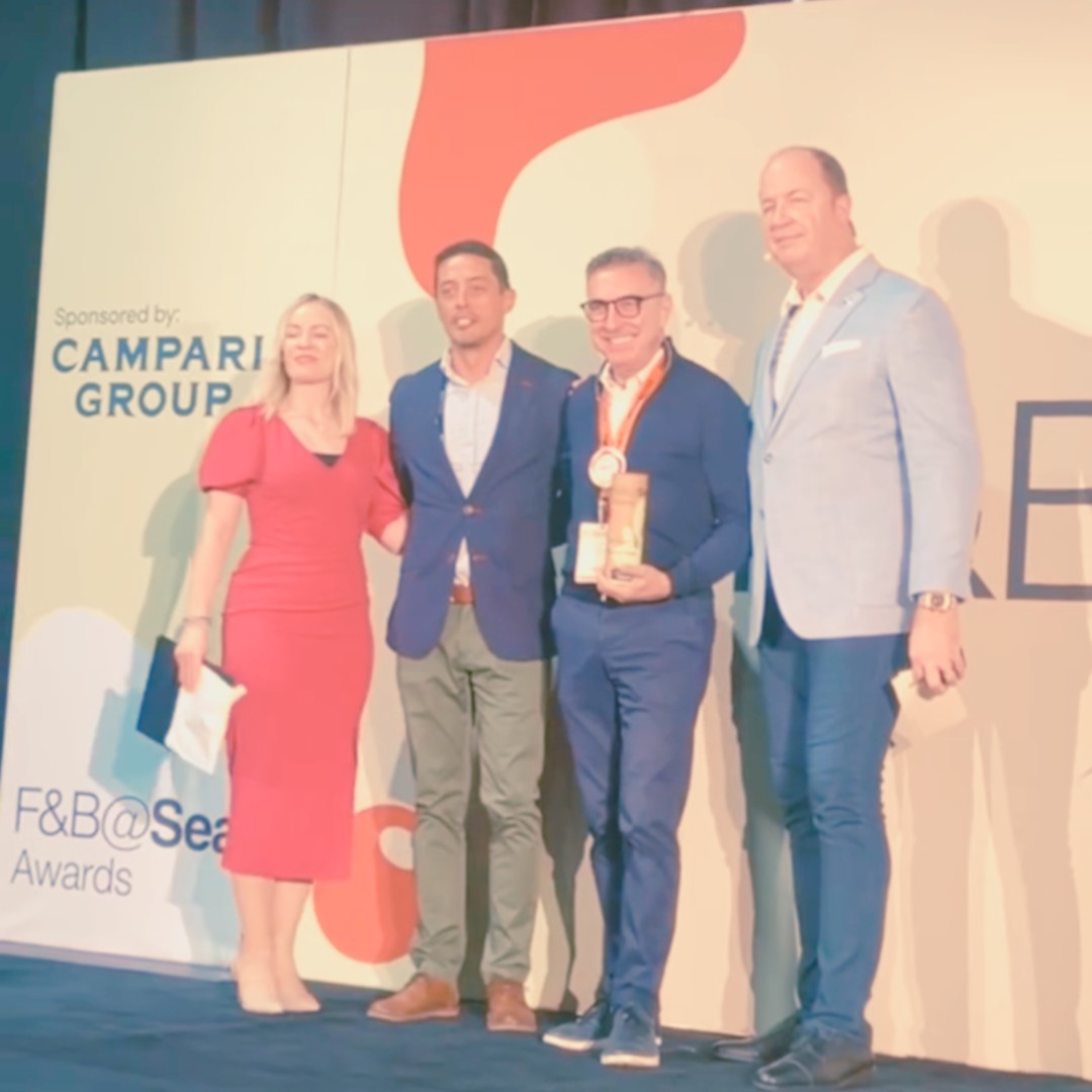 Four people stand together smiling on a convention stage for a photo opportunity at the "F&B@Sea Awards". The stage backdrop indicates the category is sponsored by "CAMPARI GROUP". The group consists of a woman in a red dress, two men in suits, and a man holding an award while wearing an orange lanyard medal.