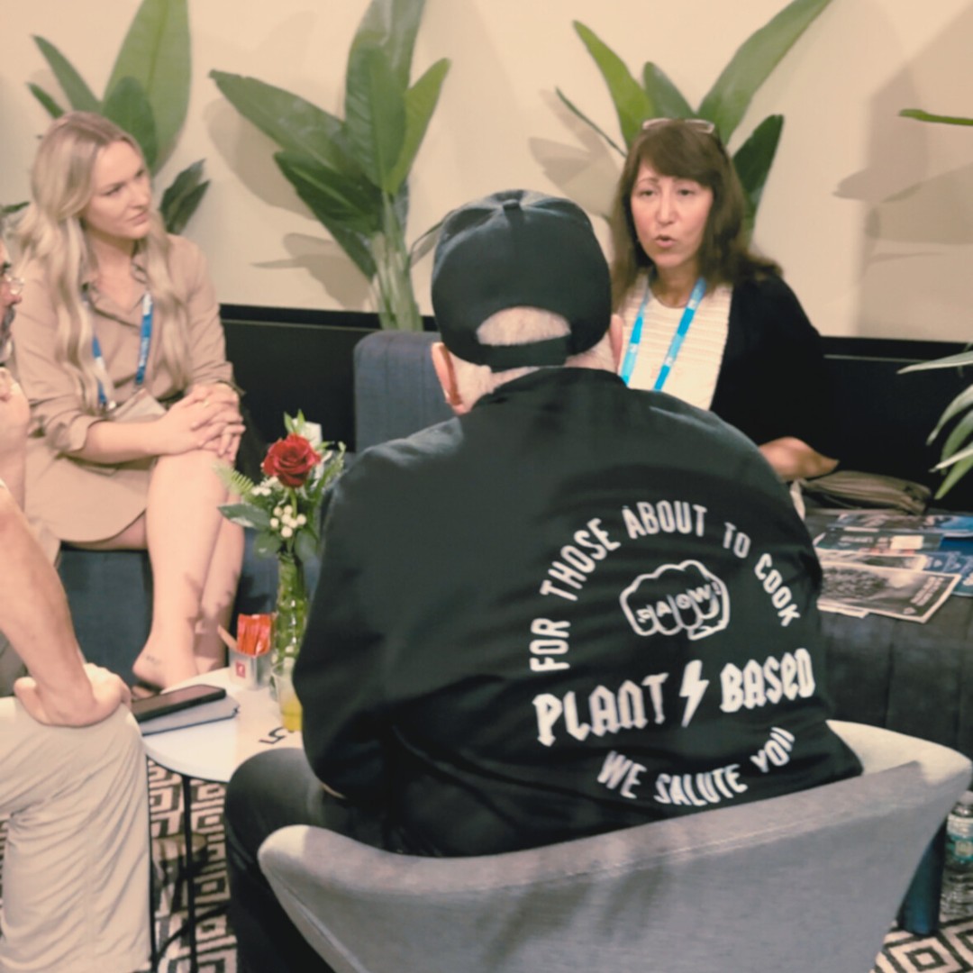 A group of people engaged in a casual meeting seated in an indoor lounge area with potted plants. In the foreground, a man sits with his back to the camera, wearing a black shirt with text that reads "FOR THOSE ABOUT TO COOK, PLANT-BASED, WE SALUTE YOU".