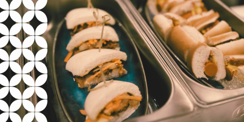 Catering trays filled with party food. The left tray holds soft white bao buns stuffed with shredded carrots and a savory filling, secured with toothpicks. The right tray contains miniature hot dogs in sliced buns. A white geometric pattern runs down the left border.