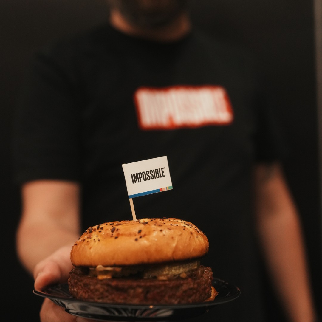 A close-up of a person holding a small black plate with a plant-based burger. A small white toothpick flag reading "IMPOSSIBLE" is stuck into the top bun. The background person wearing a black t-shirt is blurred.