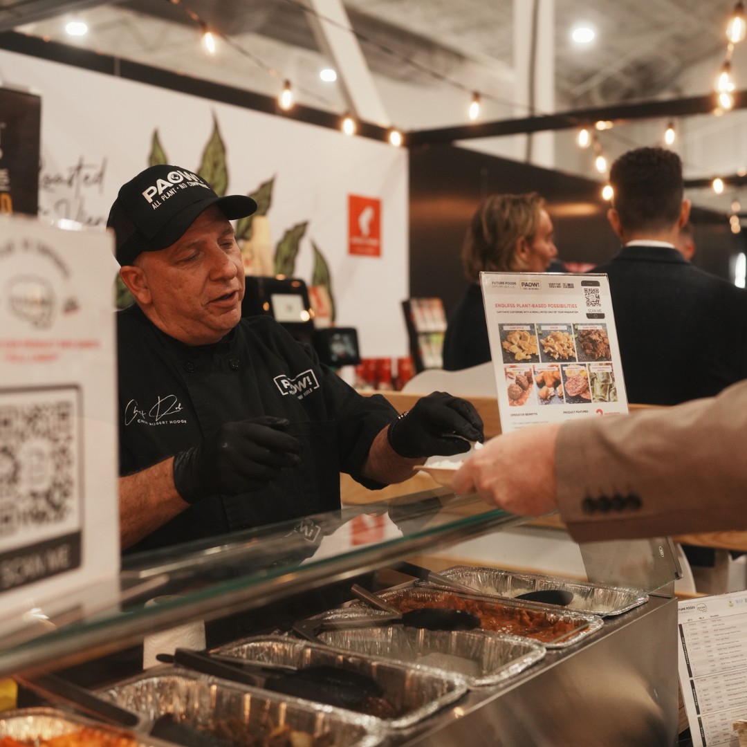 At a food expo booth, a chef in a black uniform and a "PAOW!" cap hands a small plate of food over a glass service counter to an attendee. Aluminum warming trays with food and an informational sign with a QR code are in the foreground.