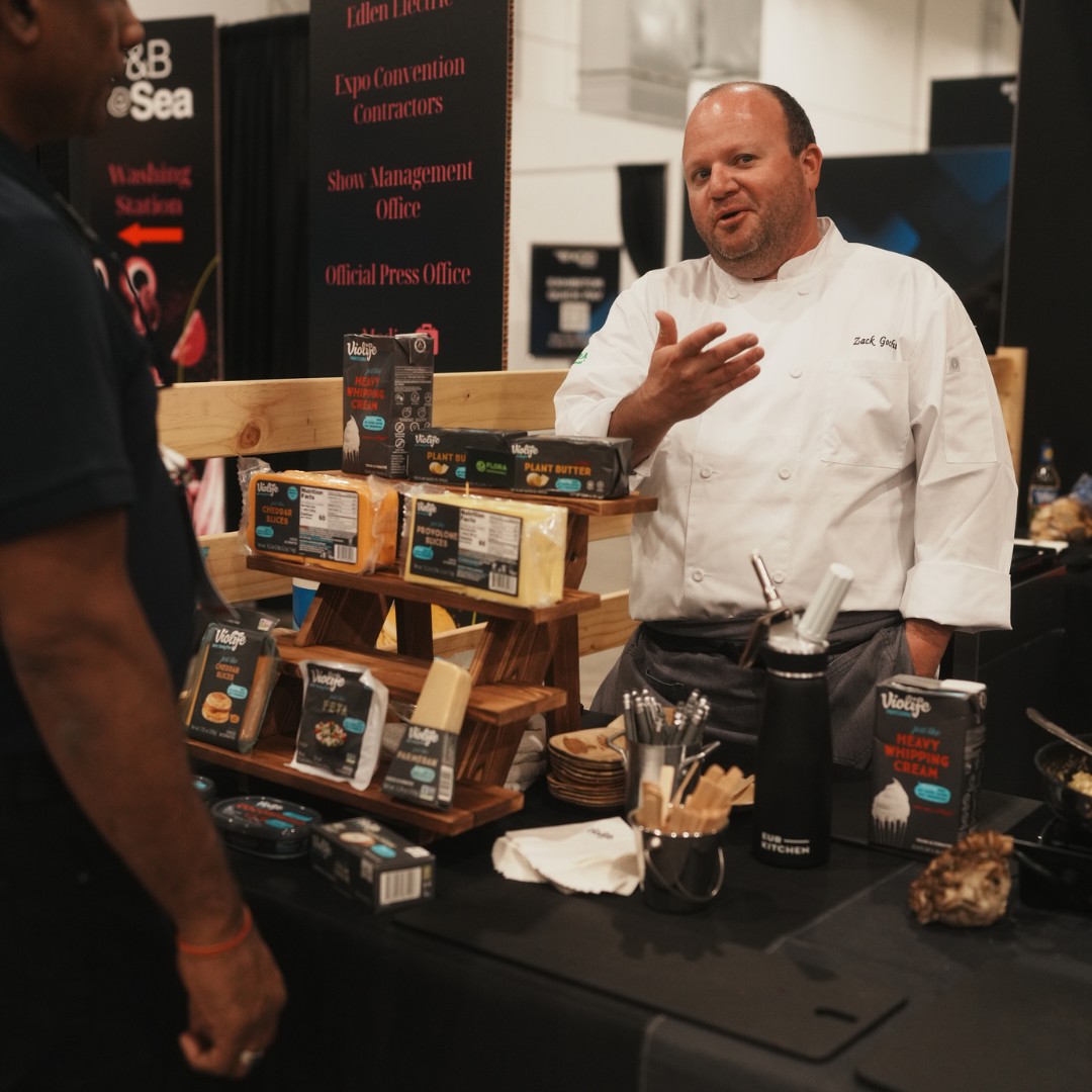 A chef in a white uniform gestures while speaking to a trade show attendee. In front of him, a variety of packaged Violife dairy-free products&mdash;including plant butter, cheddar slices, and feta&mdash;are displayed on a small tiered wooden shelf.