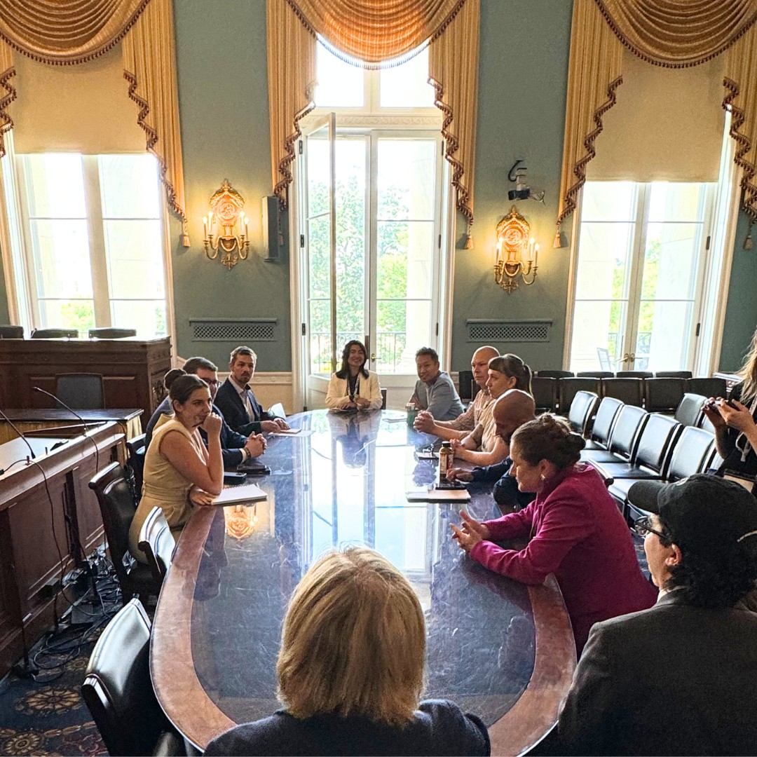 A group of professionals gathered for a meeting around a very long, reflective wooden conference table in a grand room with high ceilings, tall windows, and ornate golden drapery.