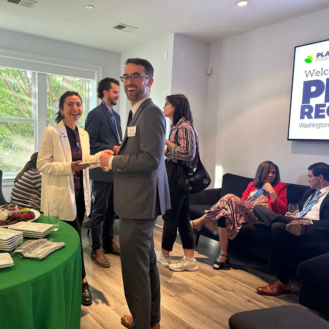 A lively indoor reception with attendees networking and laughing in a lounge area. In the foreground, a round table draped in green cloth holds plates and snacks, while a digital screen on the right wall displays a welcoming message.