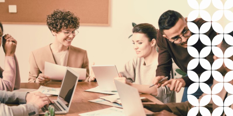 A group of diverse colleagues collaborating around a wooden conference table in a bright office. A woman with short curly hair and glasses smiles while holding papers, a woman in the center looks at a tablet, and a man wearing glasses leans over to point out details to a coworker. Laptops and notes are scattered on the table, and a white geometric pattern frames the right border.