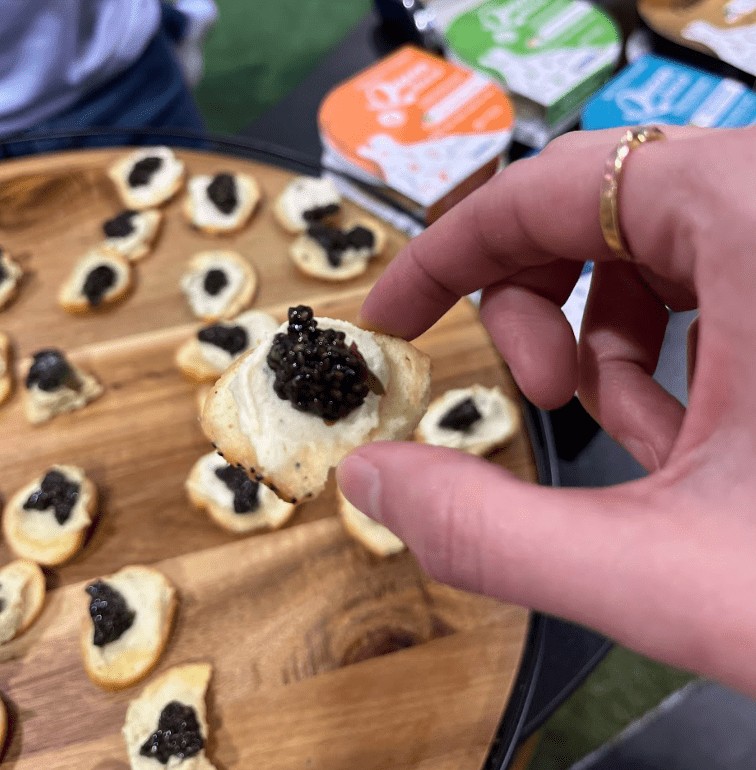 A close-up view of a person's hand holding a small piece of bread topped with a white spread and a spoonful of dark plant-based caviar over a wooden platter of similar appetizers.