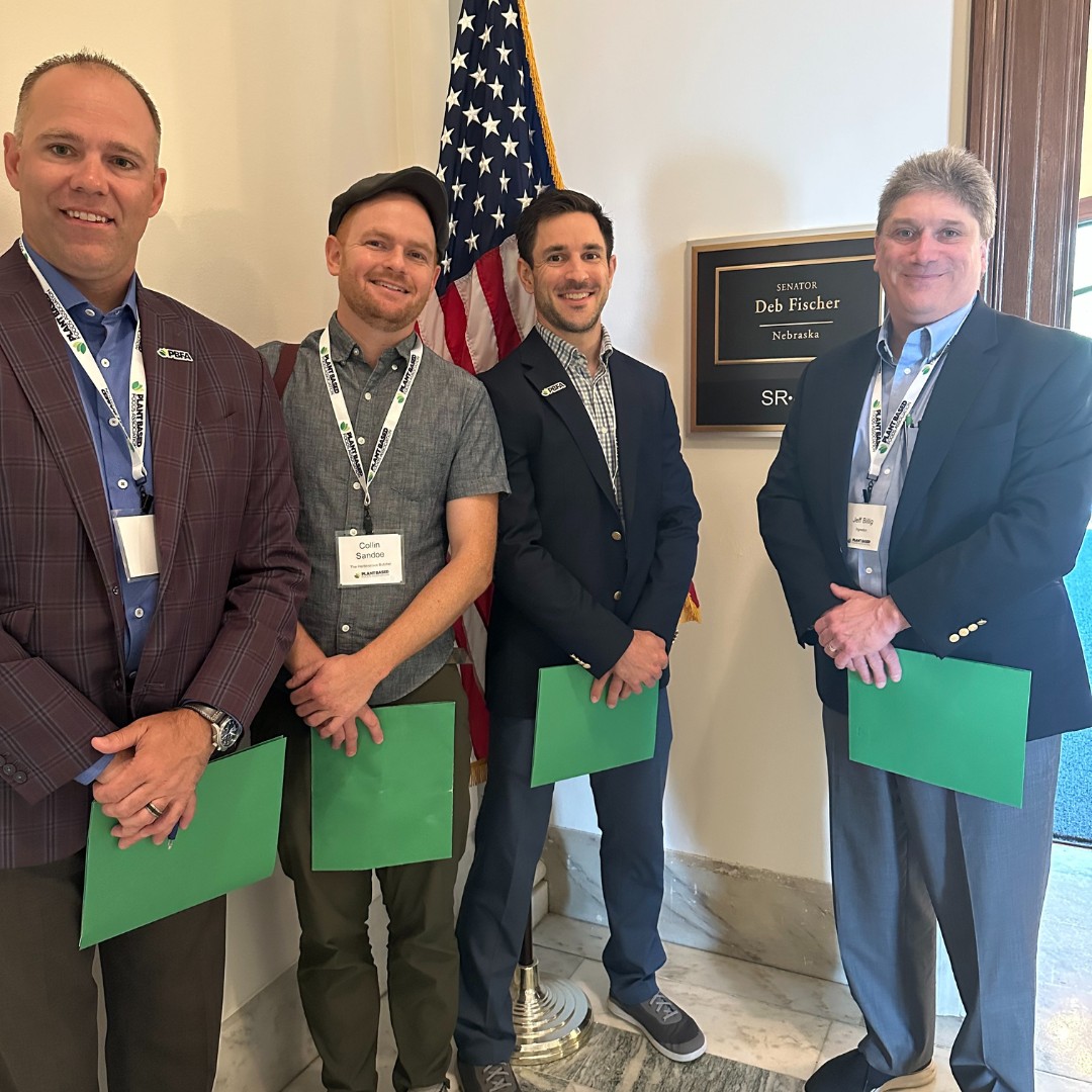 Four men wearing lanyards stand in a row next to an American flag and an office wall plaque that reads "Senator Deb Fischer Nebraska." Three of the men are holding matching green folders.