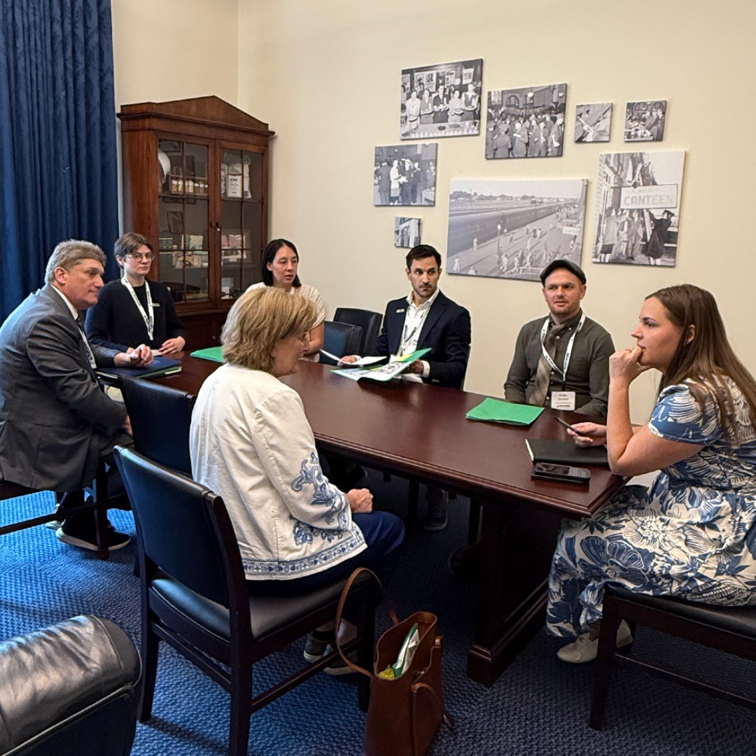 A meeting taking place inside a congressional office around a long dark wood conference table. Several advocates holding green folders are seated across from a lawmaker in a white and blue jacket. The wall behind them features several historical black-and-white photos.