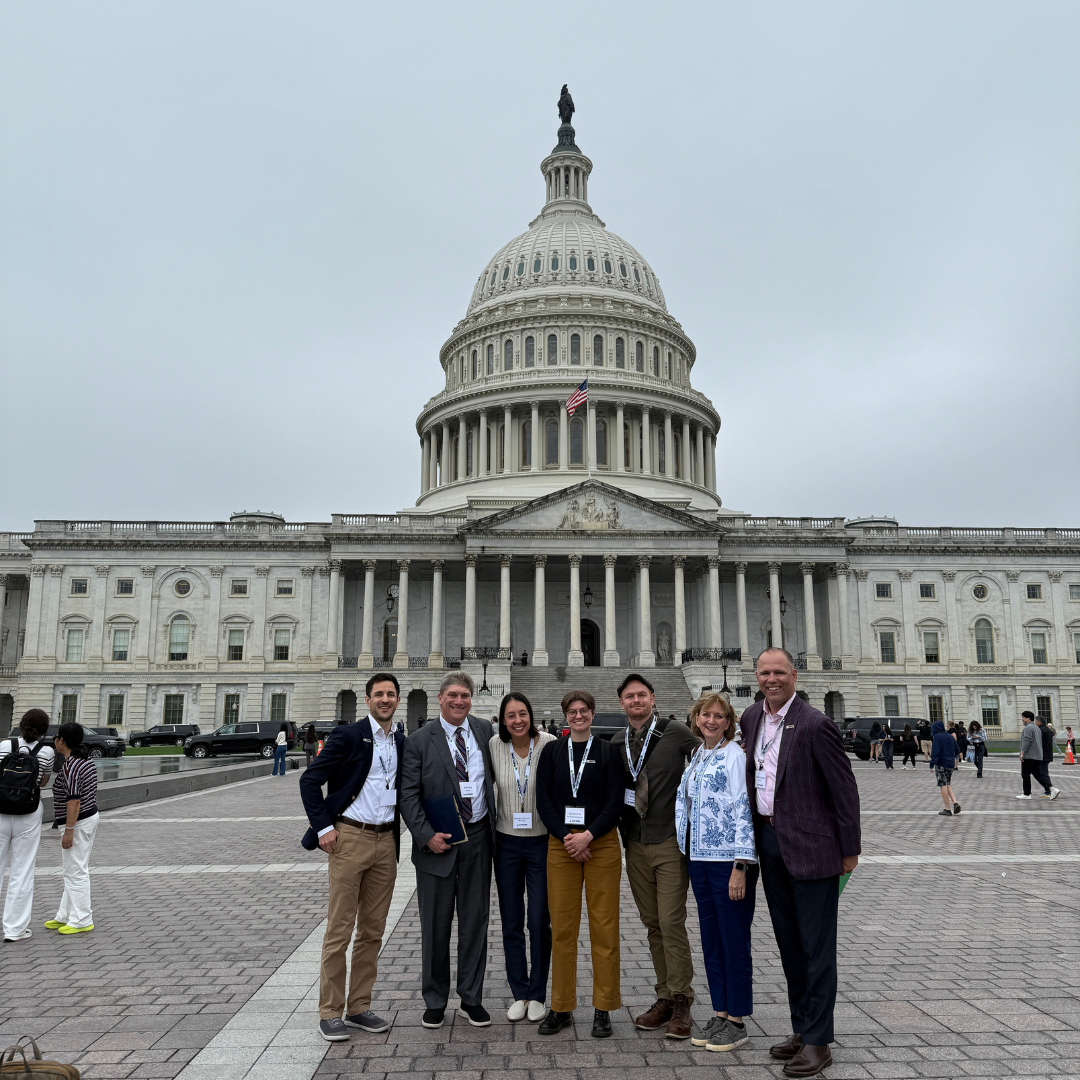 Seven advocates standing in a row on the outdoor plaza in front of the U.S. Capitol Building. They are dressed in business casual attire and suits, smiling under a cloudy sky.