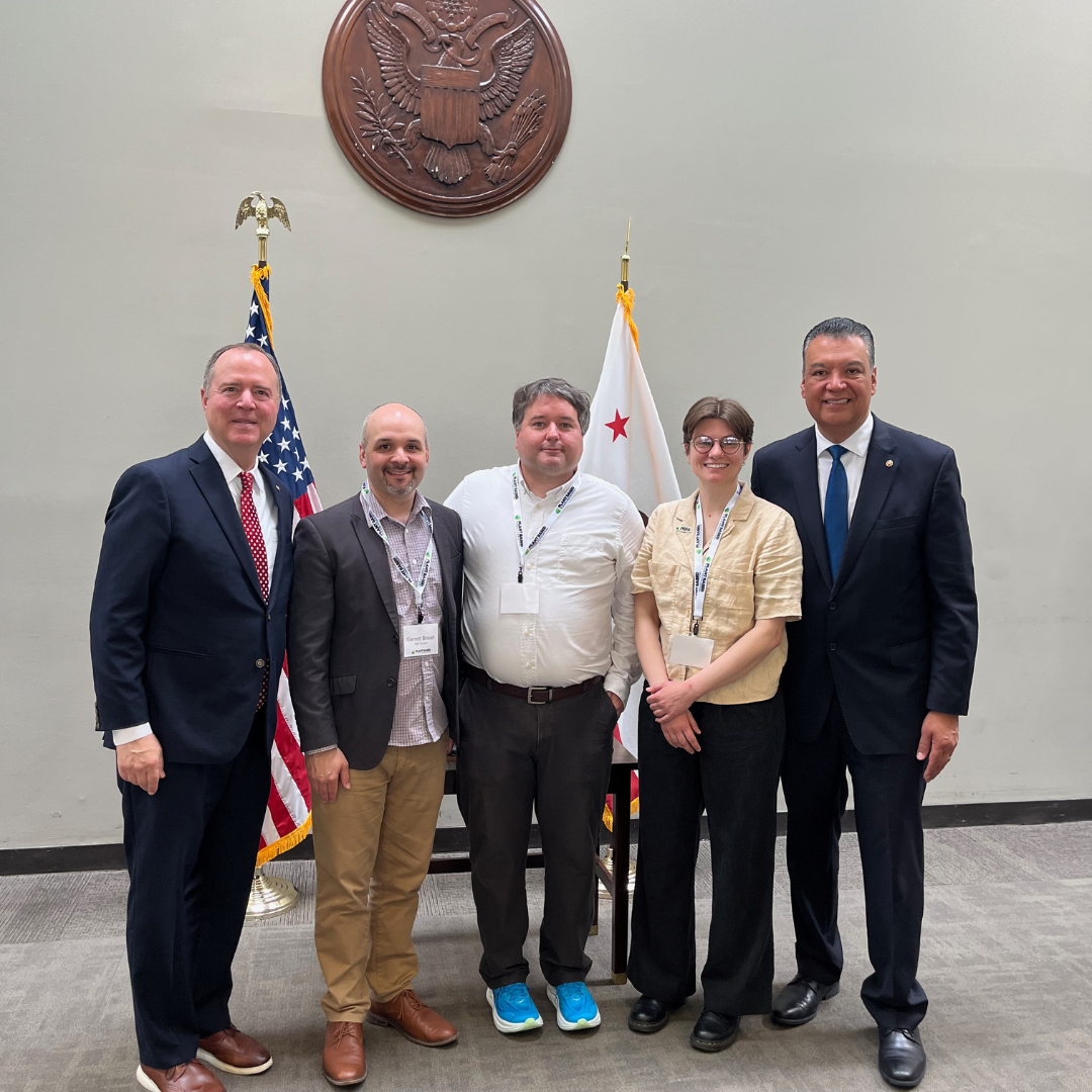Five people posing for a photo indoors against a gray wall featuring the Great Seal of the United States, an American flag, and a California state flag. The group includes Representative Adam Schiff on the far left, Senator Alex Padilla on the far right, and three advocates wearing lanyards in the center.