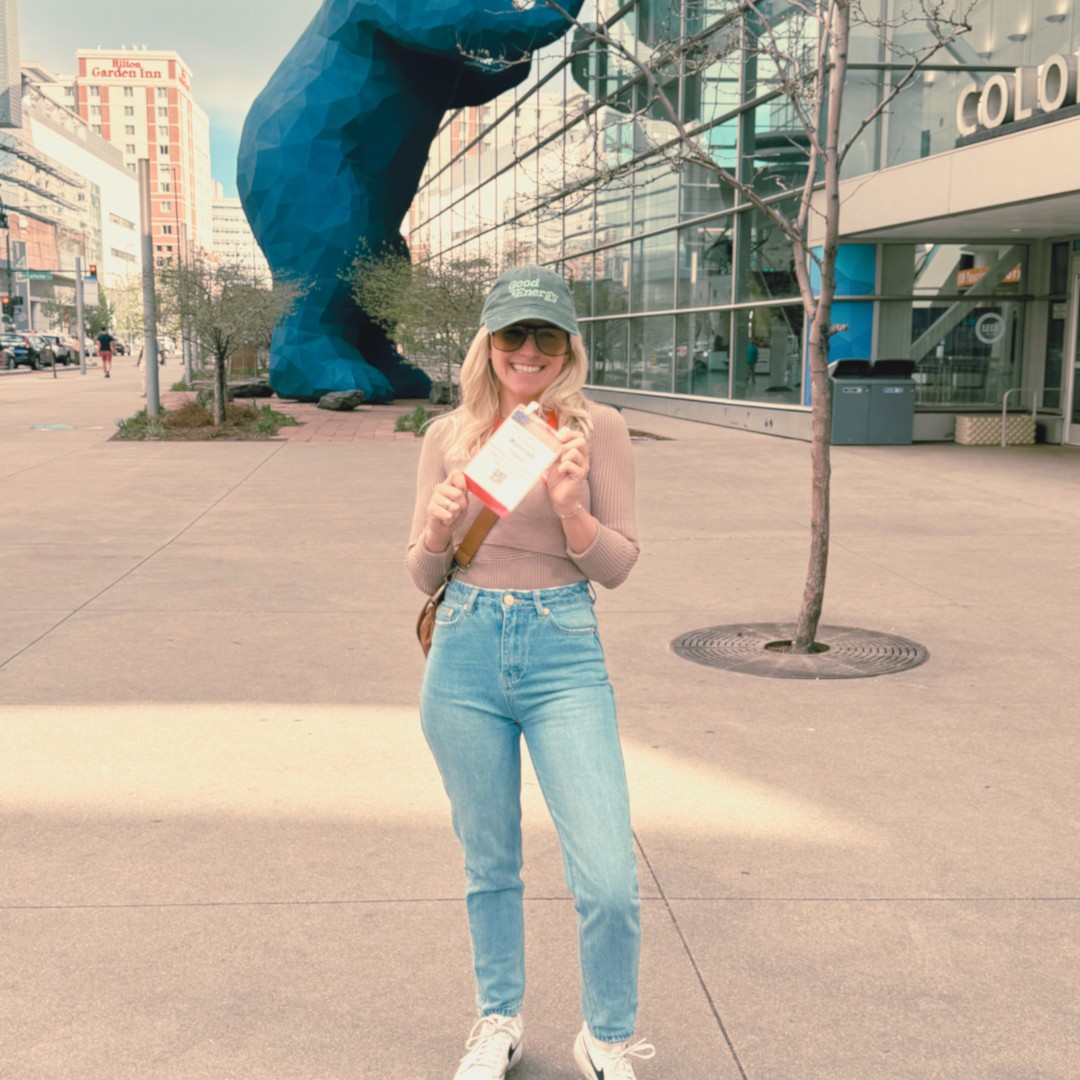 Hannah Lopez stands on a city sidewalk while holding up an event badge. A massive blue geometric bear sculpture stands in the background outside a convention building.