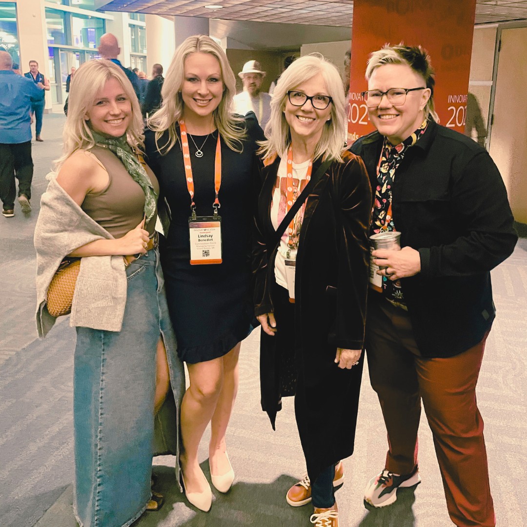 Four smiling women stand together posing for a photo at an indoor evening event. They are dressed in smart-casual attire, and two of them are wearing event badges on orange lanyards.