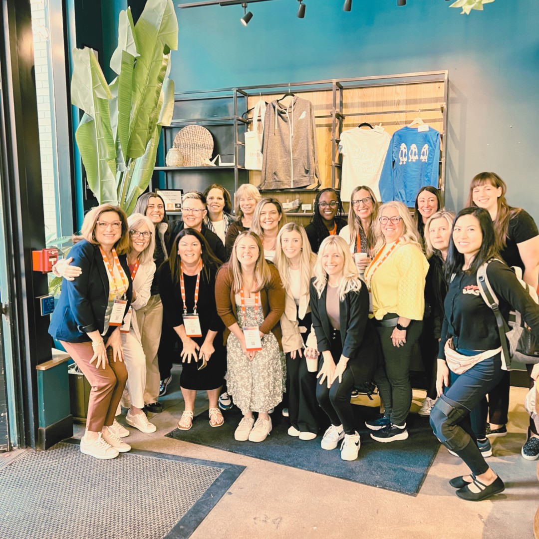 A large group of smiling women gather close together for a group photo inside a shop. The background features a teal wall and merchandise shelves holding t-shirts and hoodies.