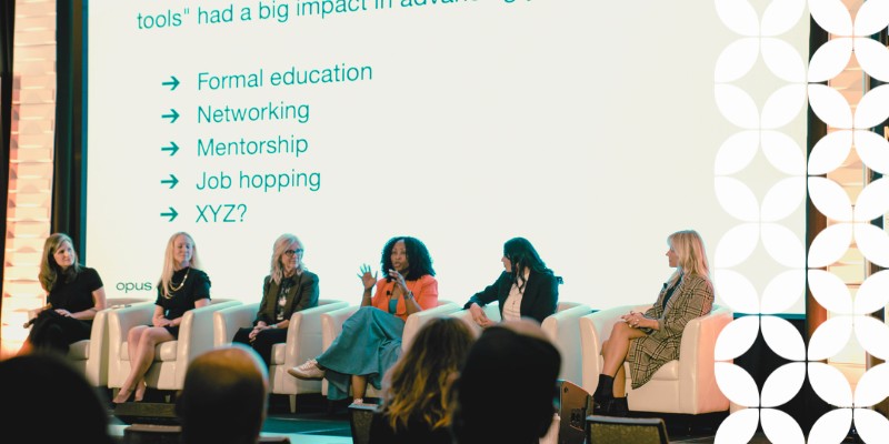 A panel presentation on a stage featuring five women seated in white leather armchairs. The speaker in the center wears an orange blazer and turquoise skirt, gesturing with her hands as she speaks. The large projection screen behind them lists tools for advancement: "Formal education", "Networking", "Mentorship", "Job hopping", and "XYZ?". A white geometric pattern frames the right edge of the image.