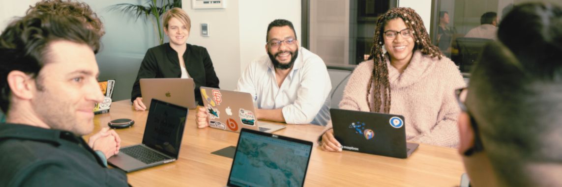 A diverse group of six people sitting around a table in a modern office, engaged and smiling, with laptops open, conveying a collaborative atmosphere.