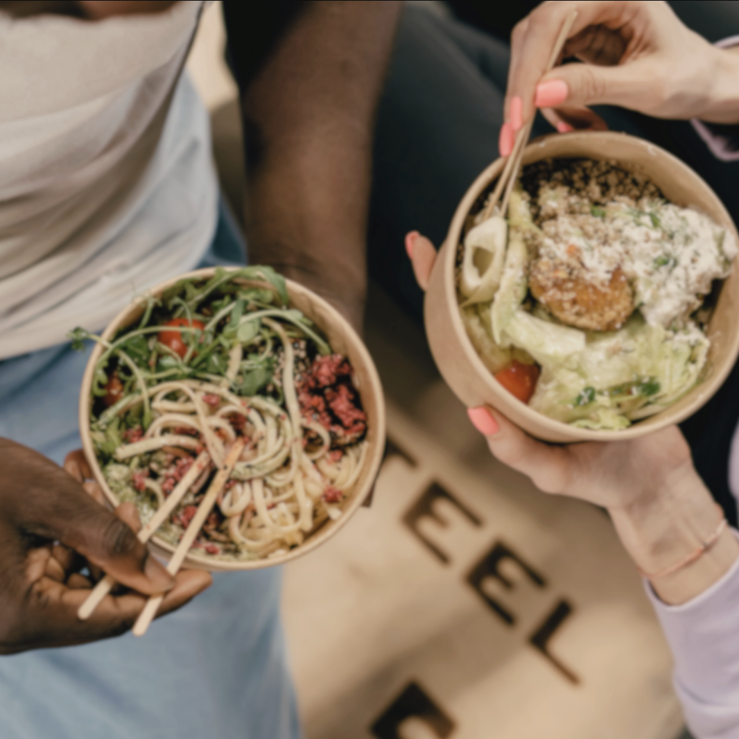 Two people holding bowls of fresh salad and pasta with chopsticks, showing diverse, healthy meals in an inviting, communal setting.
