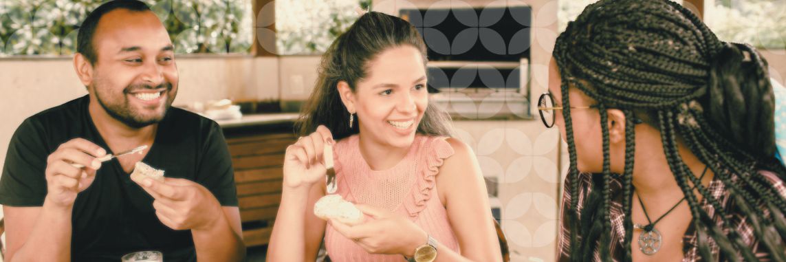 Three friends sit around a table sharing a meal together indoors, with two of them spreading butter or dip onto pieces of bread while talking and laughing. A subtle geometric pattern overlays the background.