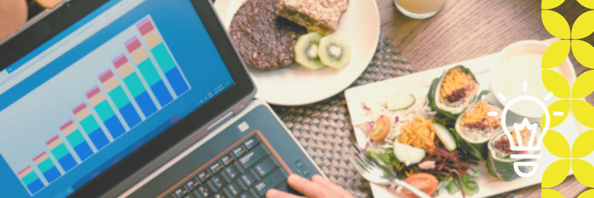 A laptop displaying a colorful bar chart sits on a table next to a plate of healthy food, including kiwi and salad. A bright, energetic atmosphere is created by the vibrant colors and creative light bulb illustration.