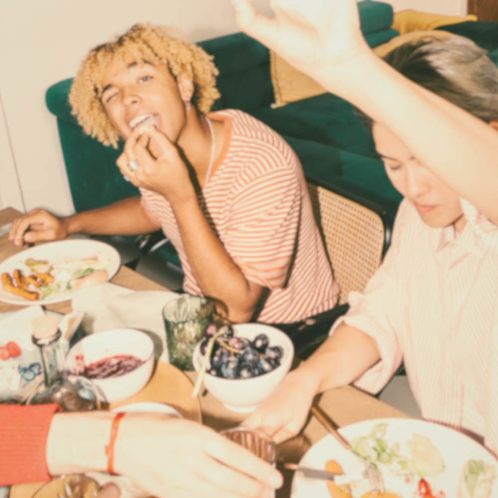 Image of two people enjoying a casual meal at a table. One person is eating, while the other raises an arm, surrounded by plates of food and a bowl of grapes.