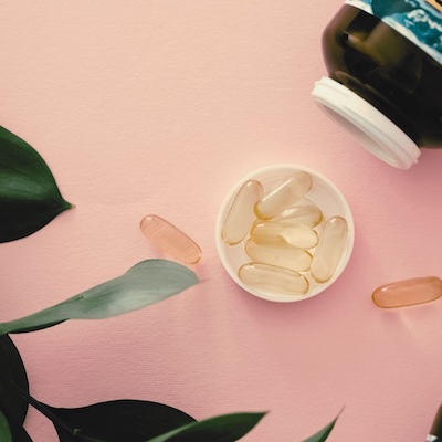 Capsules scattered on a pink surface with some inside a white cap, next to an open bottle. Green leaves frame the scene, conveying a natural feel.