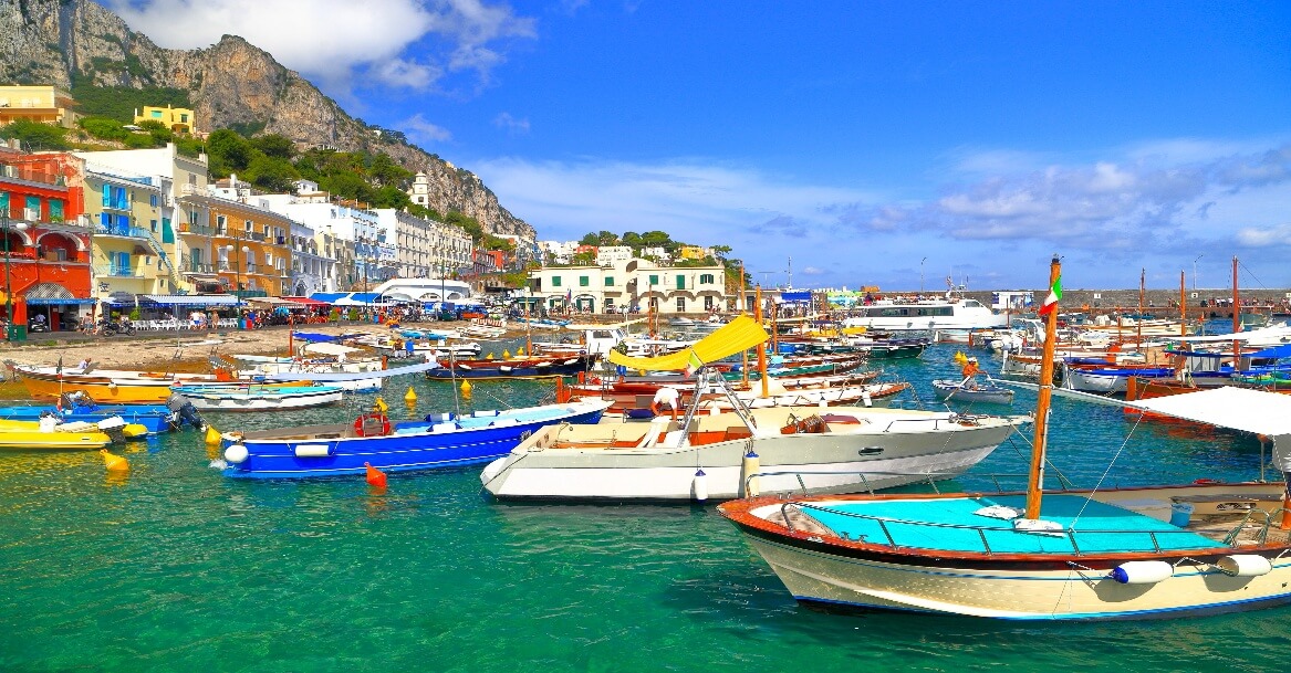 The image depicts a picturesque coastal scene with a variety of colorful boats docked in a harbor, surrounded by a charming, hilly town with vibrant buildings under a partly cloudy blue sky.AI-generated content may be incorrect.