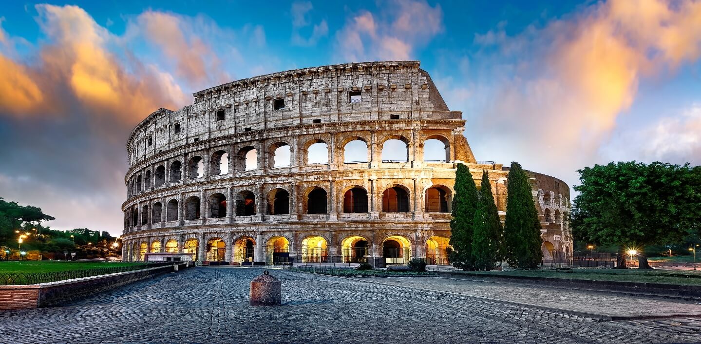 The image depicts the iconic Colosseum in Rome, illuminated against a dramatic sunset sky.AI-generated content may be incorrect.