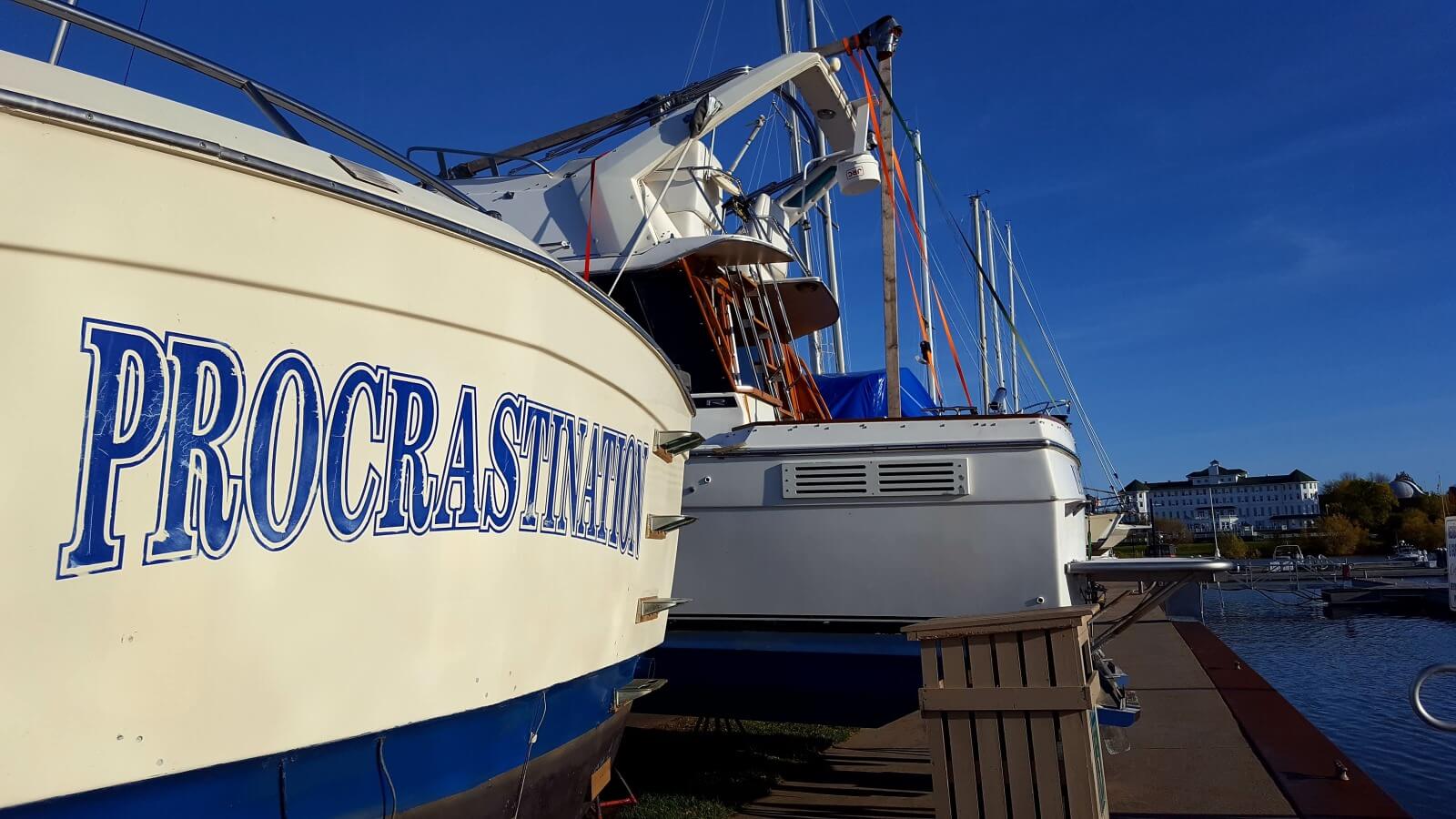 A photo of a white boat with the name "Procrastination" painted in blue letters docked at a marina with the Hotel Chequamegon, a white building with a green roof, visible in the distance.