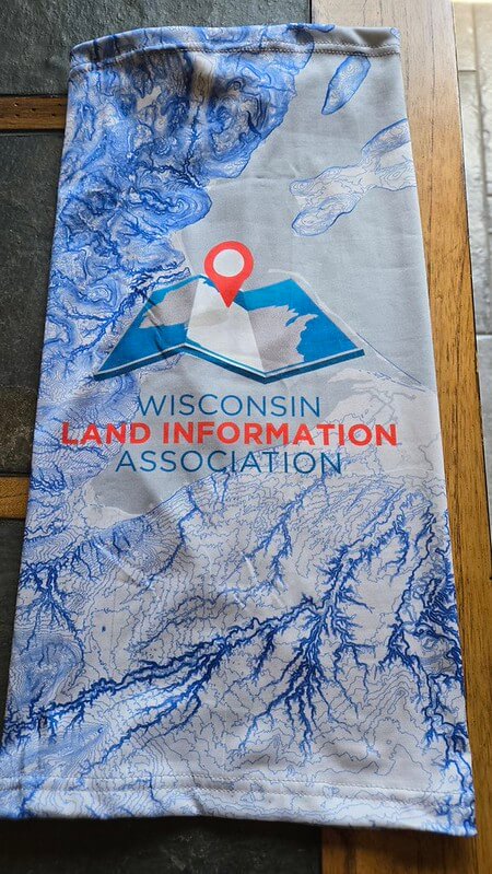 Picture of the neck gaiter sold for the WLIA 2026 spring conference.  Gaiter has images of  blue elevation contours of Ashland Area, along with the WLIA logo
